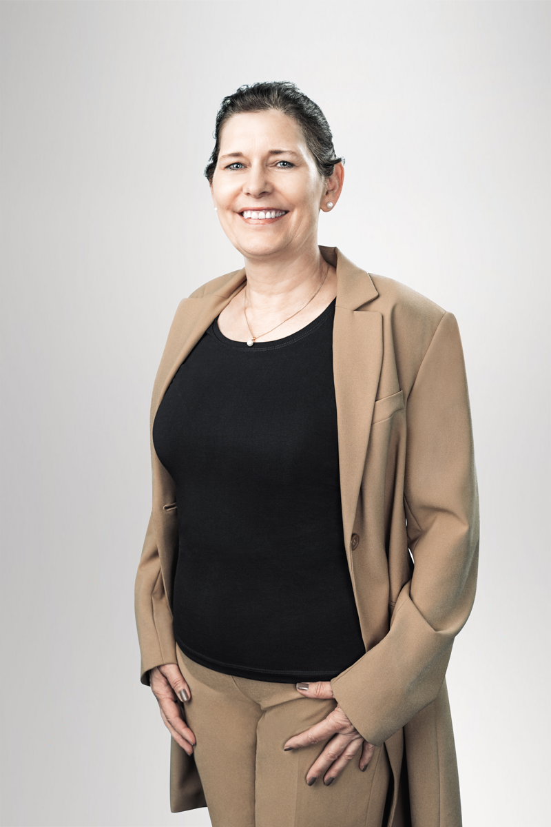 Eine lächelnde Frau in einem zweitönigen Outfit, bestehend aus einem schwarzen Top und einem beigen Blazer, steht vor einem neutralen Hintergrund und posiert mit verschränkten Fingern auf Höhe ihrer Taille.