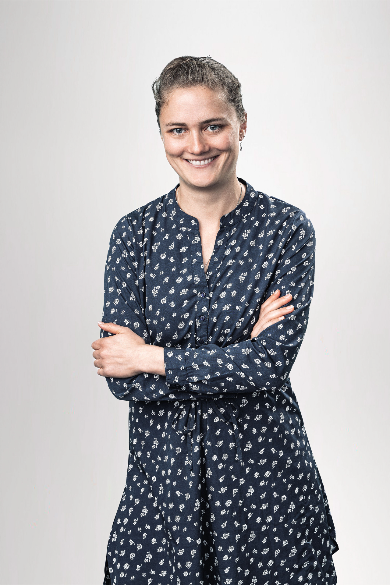 Eine lächelnde Frau steht mit verschränkten Armen vor einem neutralen Hintergrund und trägt ein langärmliges, gemustertes Kleid in Dunkelblau.