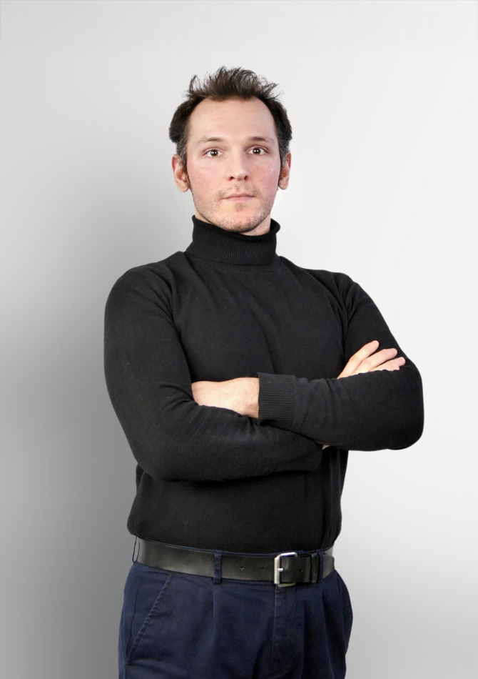 Ein junger Mann mit kurzem, dunklem Haar steht mit verschränkten Armen vor einem hellen, unverzierten Hintergrund. Er trägt einen schwarzen Rollkragenpullover und eine dunkle Hose.