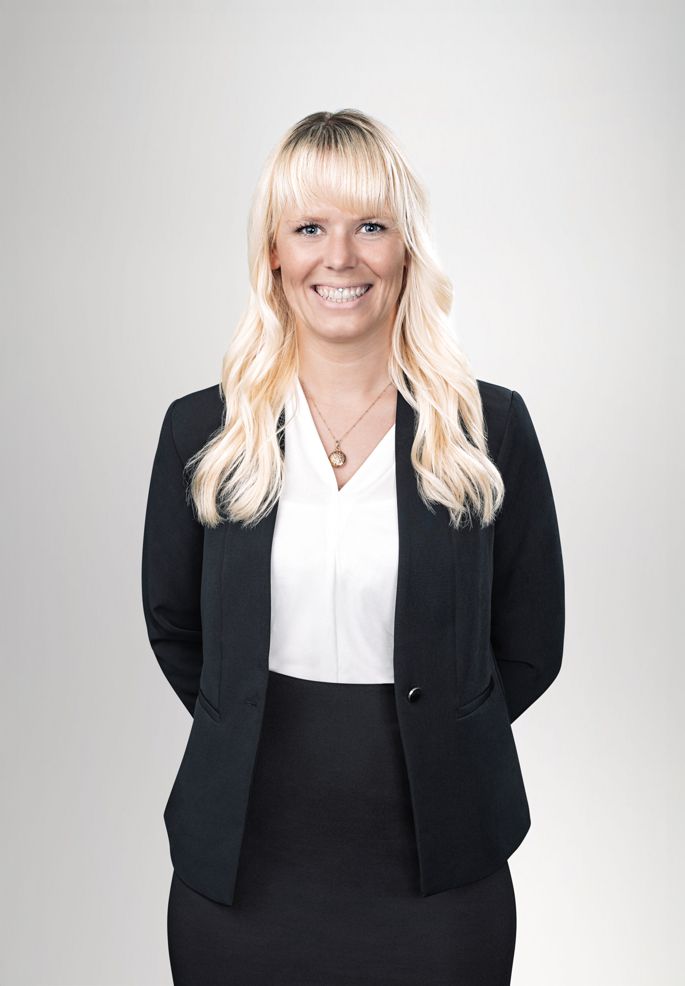 Eine lächelnde Frau mit langen blonden Haaren trägt ein schwarzes Blazerjackett und ein weißes Oberteil. Sie steht mit geschlossenen Händen vor ihrem Körper und hat einen neutralen hellen Hintergrund.