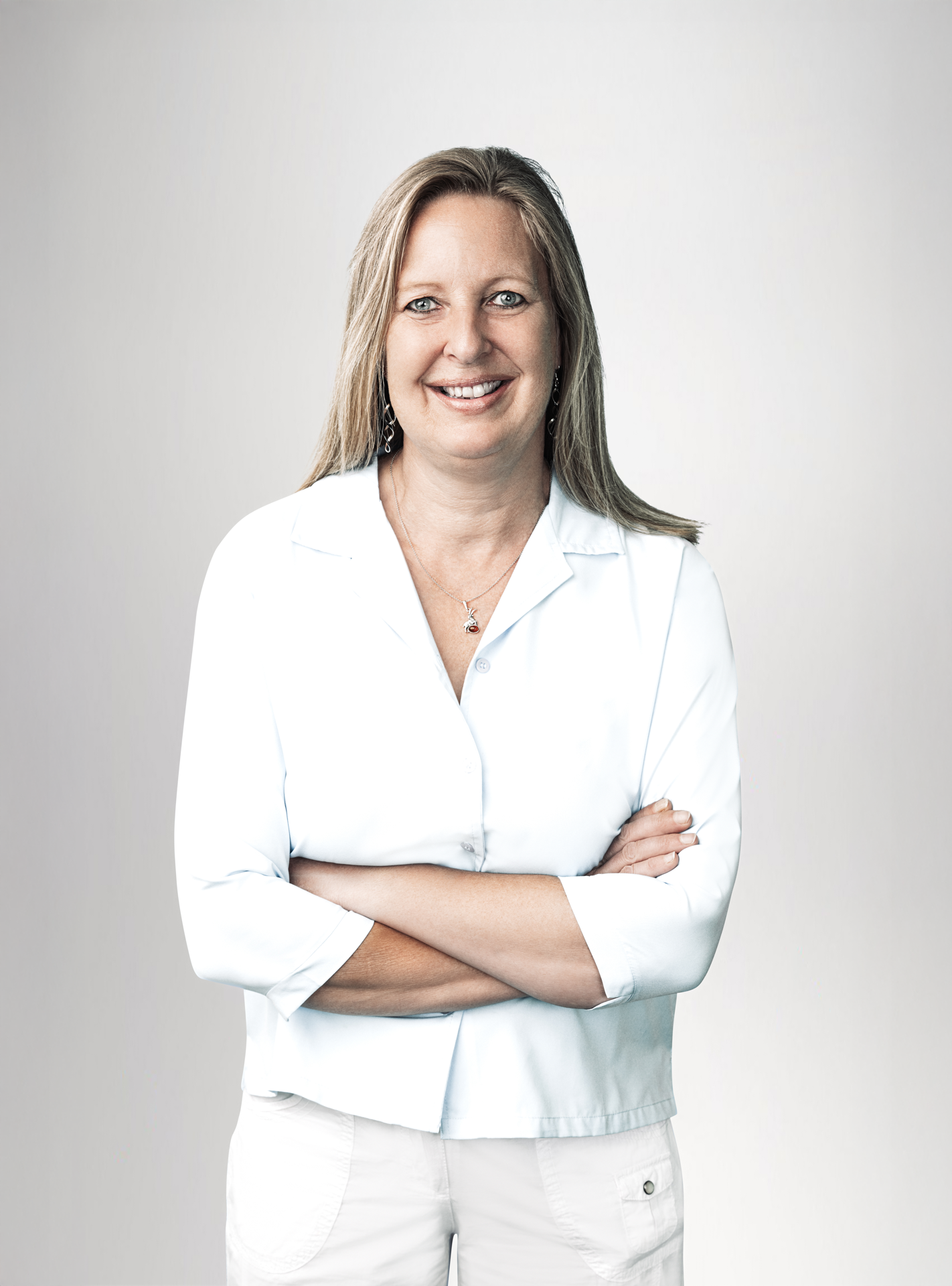 Eine lächelnde Frau mit langen, blonden Haaren steht mit verschränkten Armen vor einem hellen Hintergrund. Sie trägt ein hellblaues Oberteil und hat Ohrringe in den Ohren. Ihre Pose strahlt Selbstbewusstsein aus.