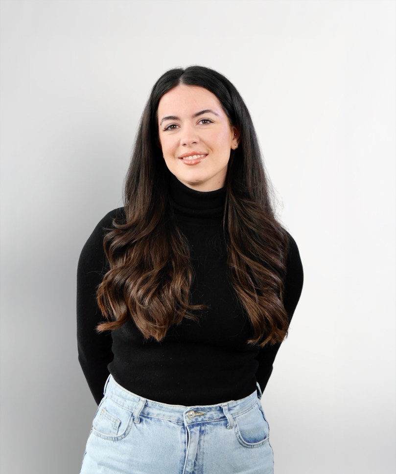 Eine Frau mit langen, lockigen, dunklen Haaren trägt einen schwarzen Rollkragenpullover und helle Jeans, während sie lächelt und die Hände hinter dem Rücken verschränkt.