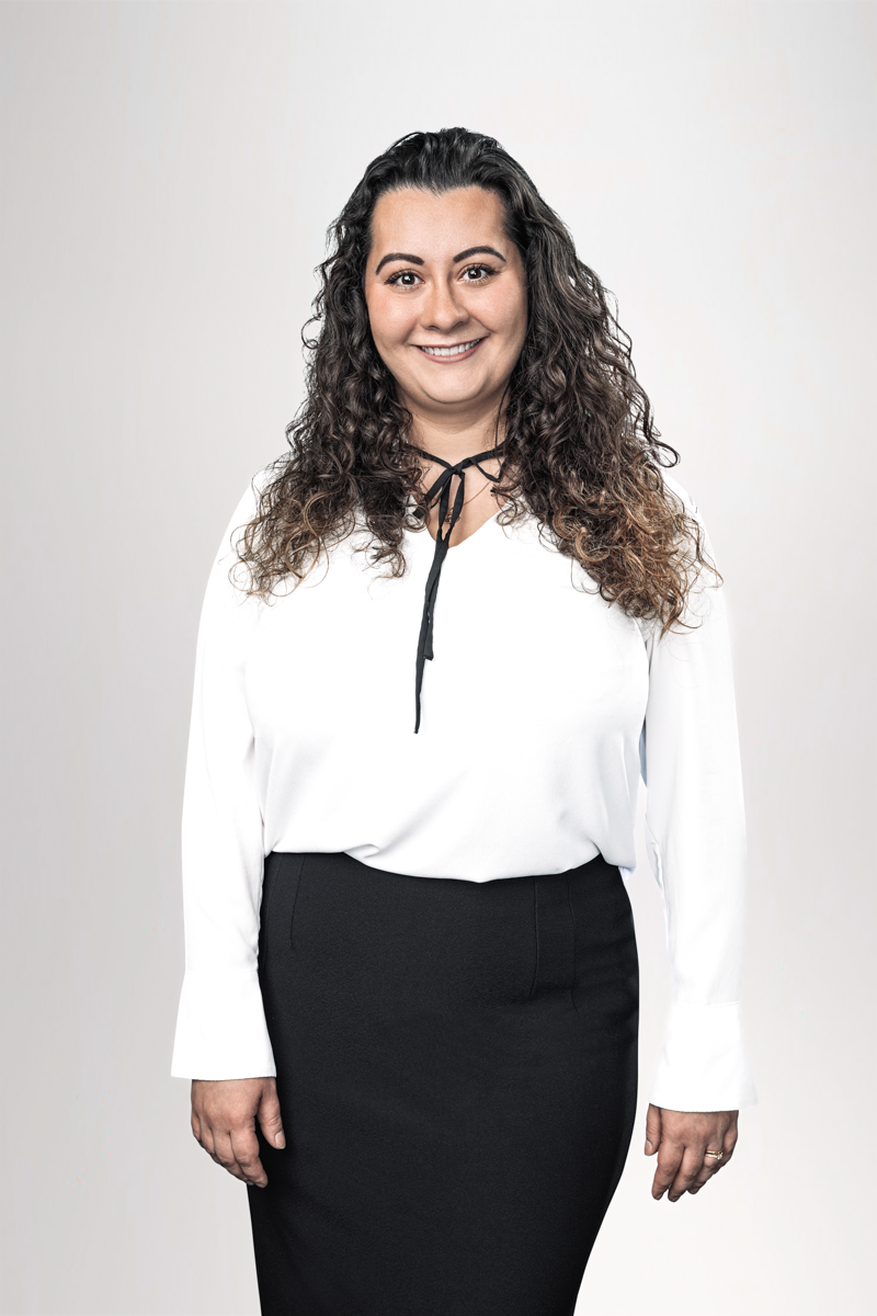 Eine lächelnde Frau mit lockigem, langen Haar trägt eine weiße Bluse mit einem schwarzen Krawattenband und einen schwarzen Rock. Der Hintergrund ist hell und neutral.