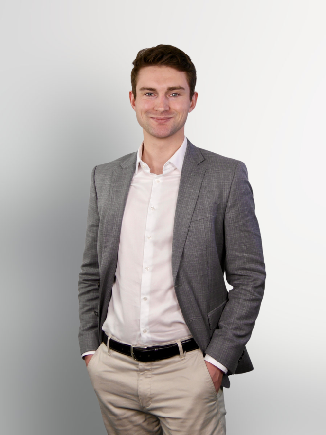 Ein junger Mann in einem grauen Blazer und heller Hemd steht mit verschränkten Armen vor einem hellen, einfarbigen Hintergrund und lächelt.