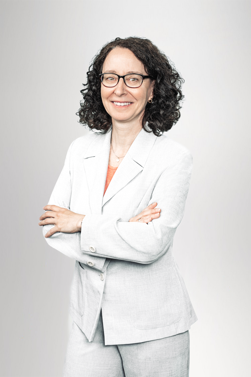 Eine lächelnde Frau mit lockigem, dunkelbraunem Haar und Brillen steht mit verschränkten Armen in einem hellen Anzug vor einem neutralen Hintergrund. Sie wirkt professionell und freundlich.