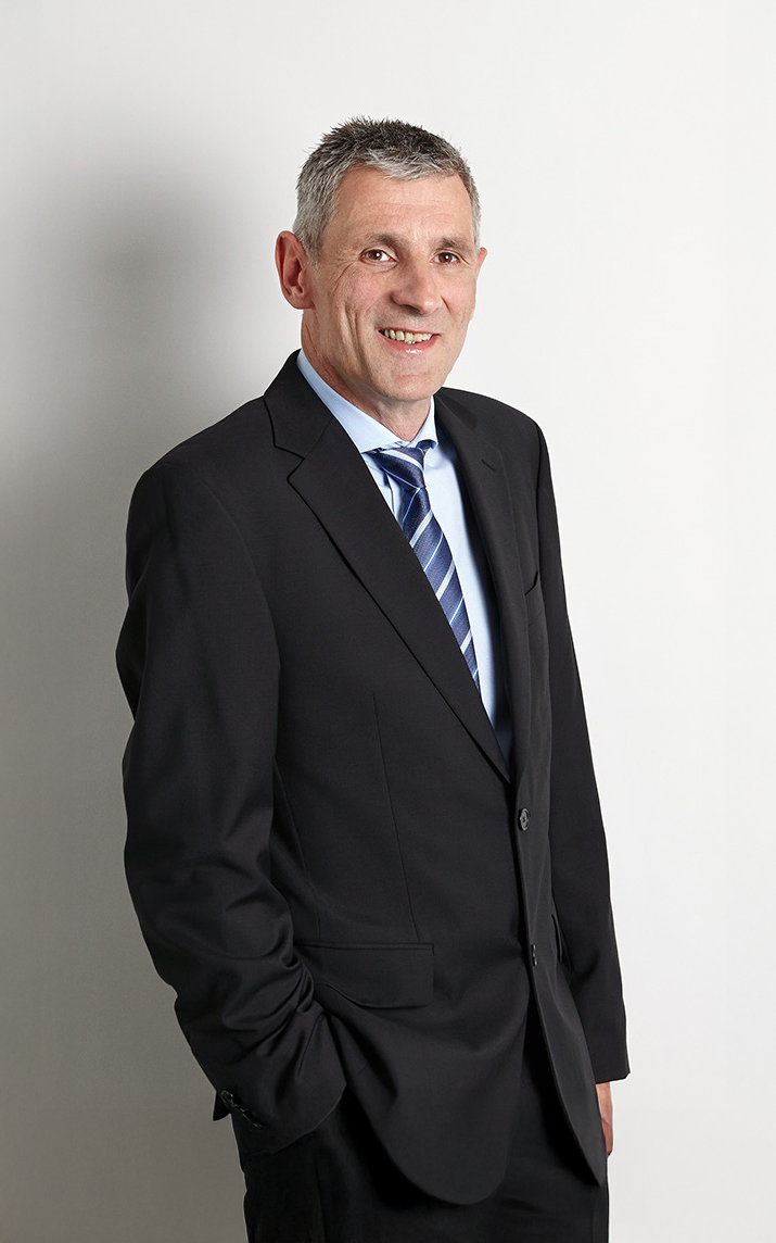 Ein Mann steht in einem Anzug mit Krawatte und lächelt freundlich, während er vor einem schlichten, hellen Hintergrund posiert.