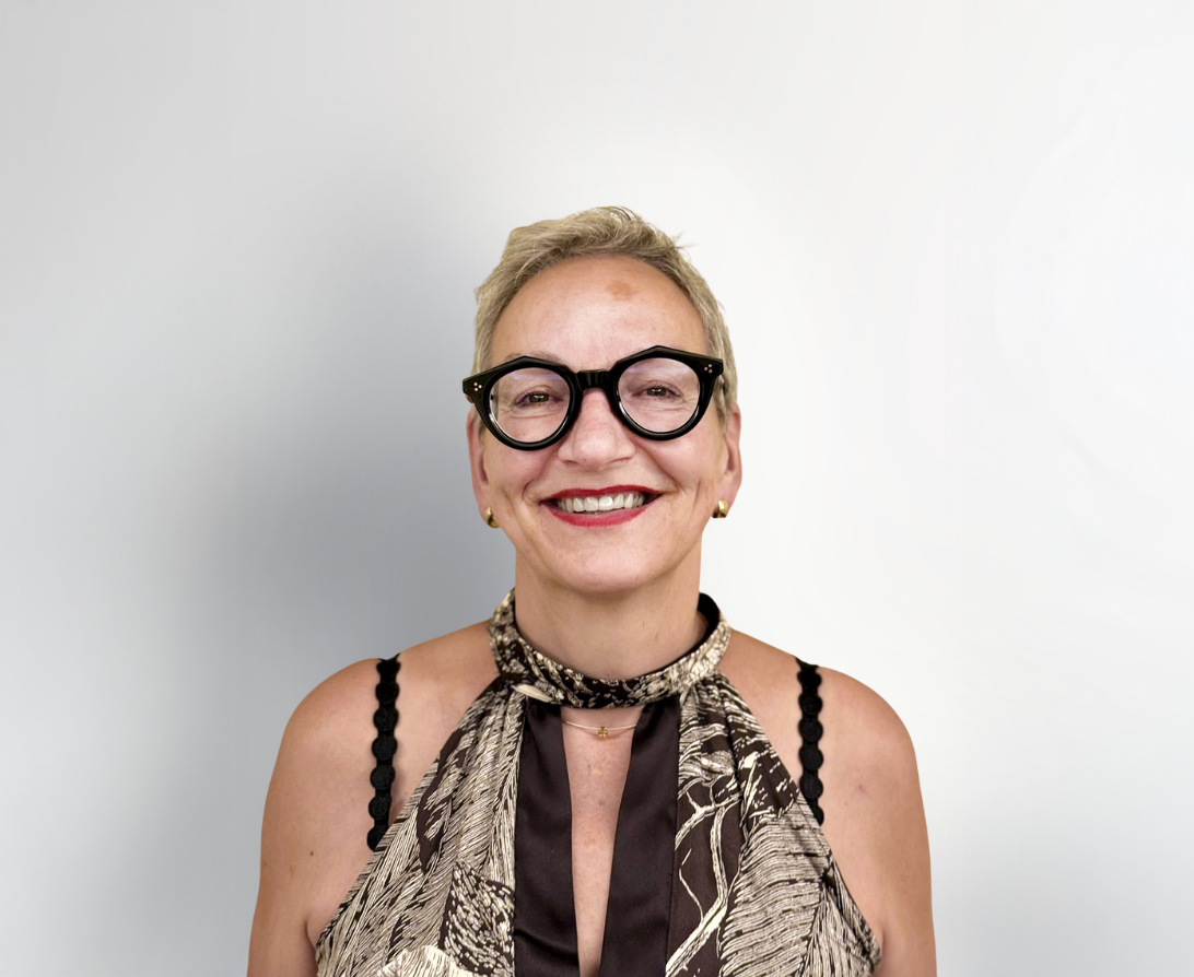 Eine lächelnde Frau mit kurzen, blonden Haaren trägt eine elegante Bluse mit einem auffälligen Muster und große, schwarze Brillen. Der Hintergrund ist hell und neutral.