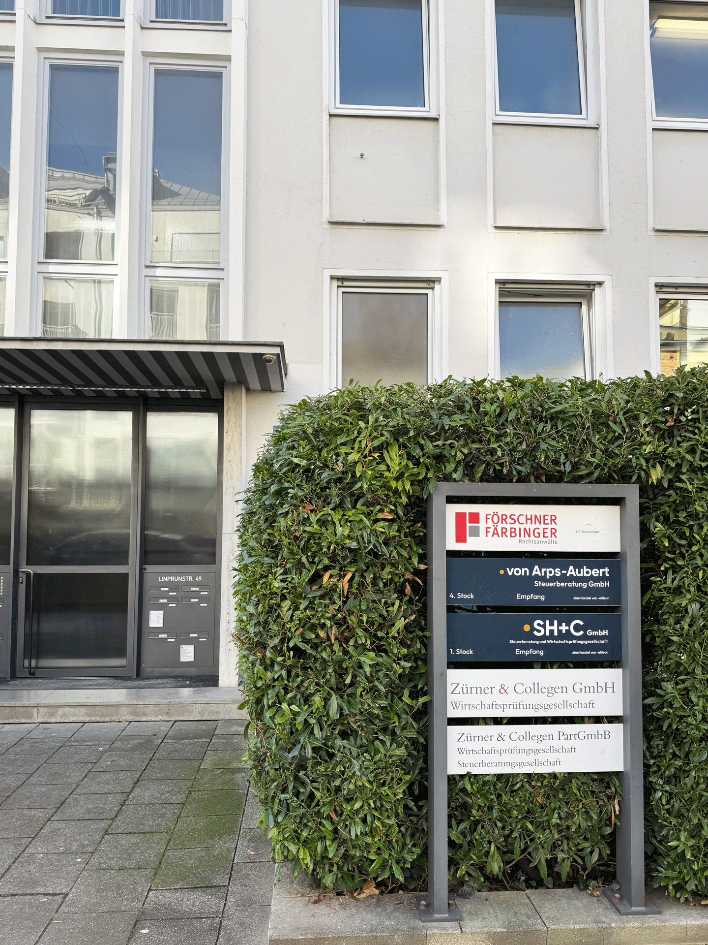 Das Bild zeigt ein modernes Gebäude mit einer Glasfront und einem Empfangsbereich sowie einem Pflanzenbeet im Vordergrund. An einem Schilderständer sind mehrere Firmennamen und ihre Kontaktdaten angezeigt, darunter "Förschner Farbinger", "von Arps-Aubert", "SH+C" und "Zirner & Collegen GmbH".