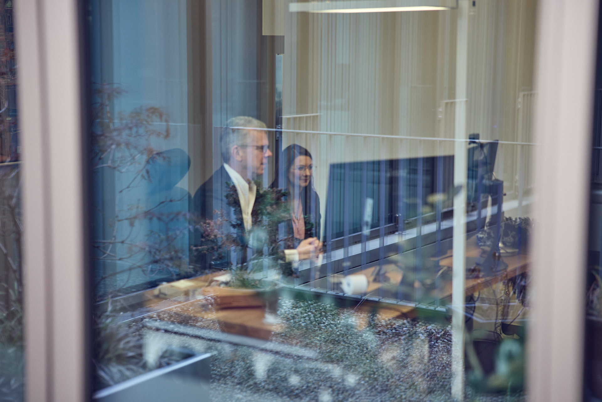 Zwei Personen sitzen an einem Tisch vor einem Computer in einem modernen Büro, während sie durch ein Fenster beobachtet werden. Im Vordergrund sind Pflanzen und der Büroausblick sichtbar.