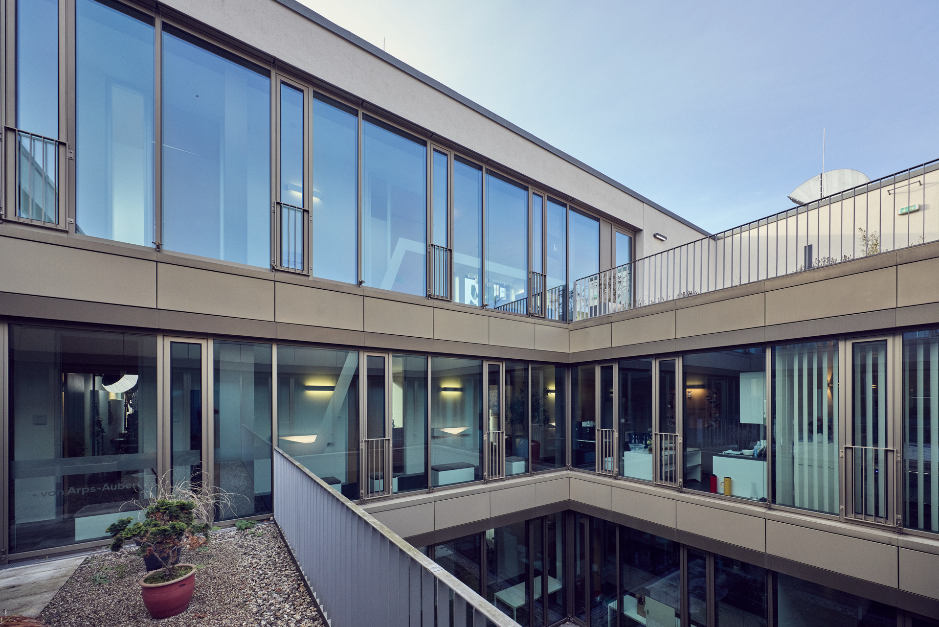 Moderne Architektur mit großen Fensterflächen und einem Innenhof, der von mehreren Etagen umgeben ist. Im Vordergrund sind kleine Pflanzen und ein Kiesbereich zu sehen.