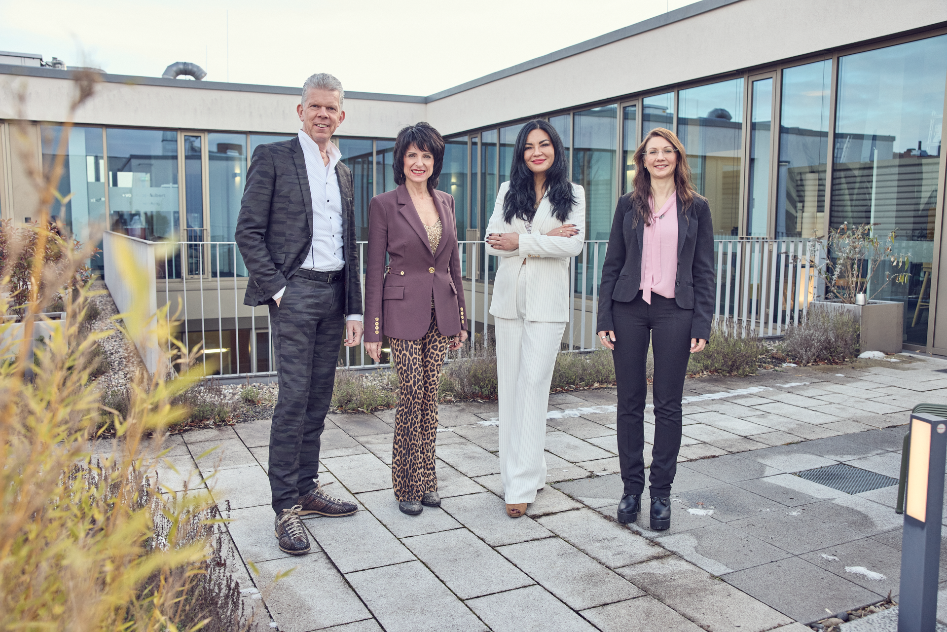 Vier Personen stehen auf einer Dachterrasse vor modernen Gebäuden. Eine Frau trägt einen braunen Blazer und einen Rock mit Tiermuster, eine andere Frau trägt einen weißen Anzug, ein Mann hat ein schwarzes kariertes Sakko an und die letzte Frau trägt eine schwarze Bluse mit einer blassroten Bluse darunter. Alle Personen lächeln in die Kamera.