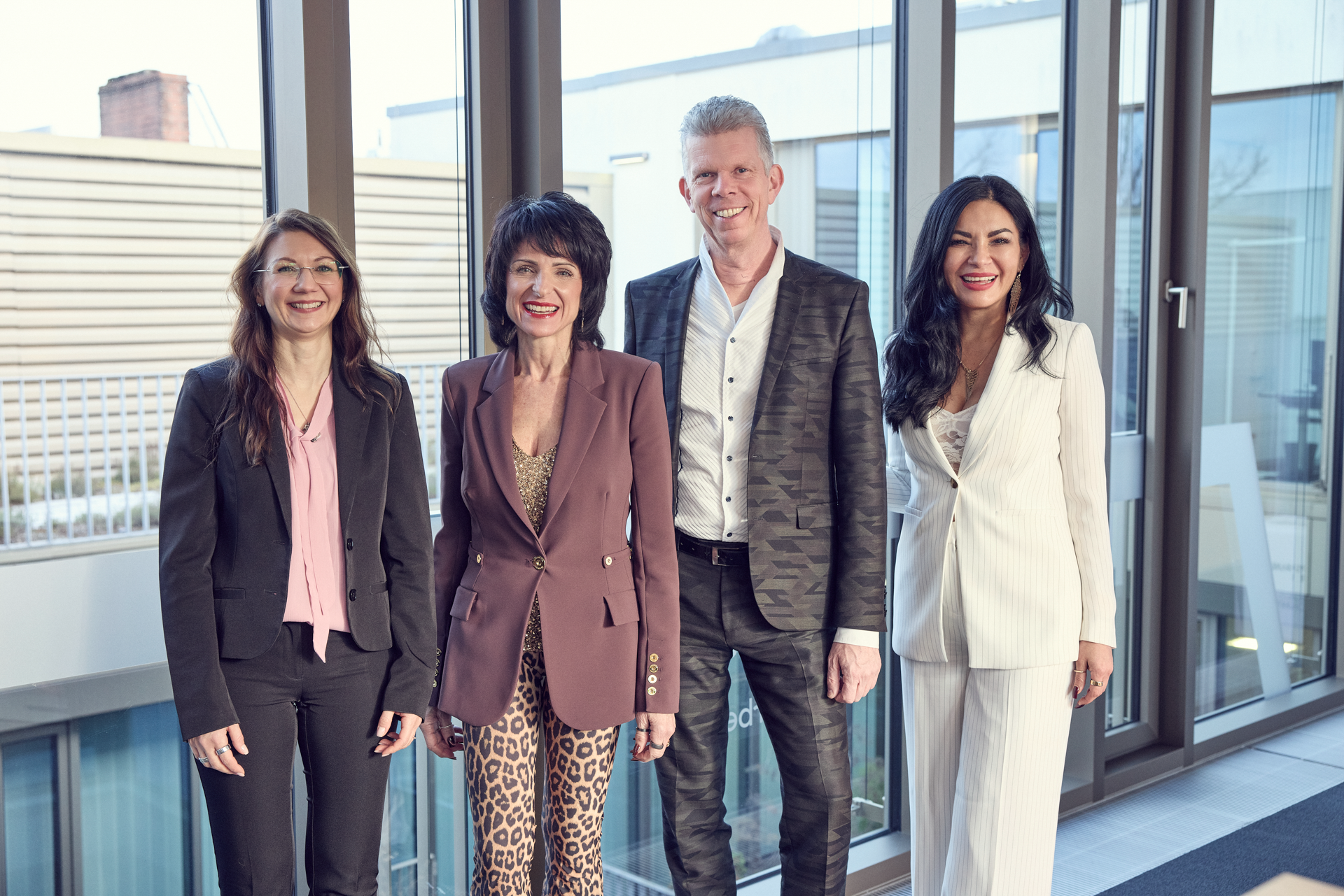 Vier Personen stehen in einem modernen, hellen Raum mit großen Fenstern. Sie posieren lächelnd nebeneinander. Alle tragen elegante, geschäftliche Kleidung: zwei Frauen in Blazern und ein Mann in einem gemusterten Anzug. Der Hintergrund zeigt eine minimalistische Architektur.
