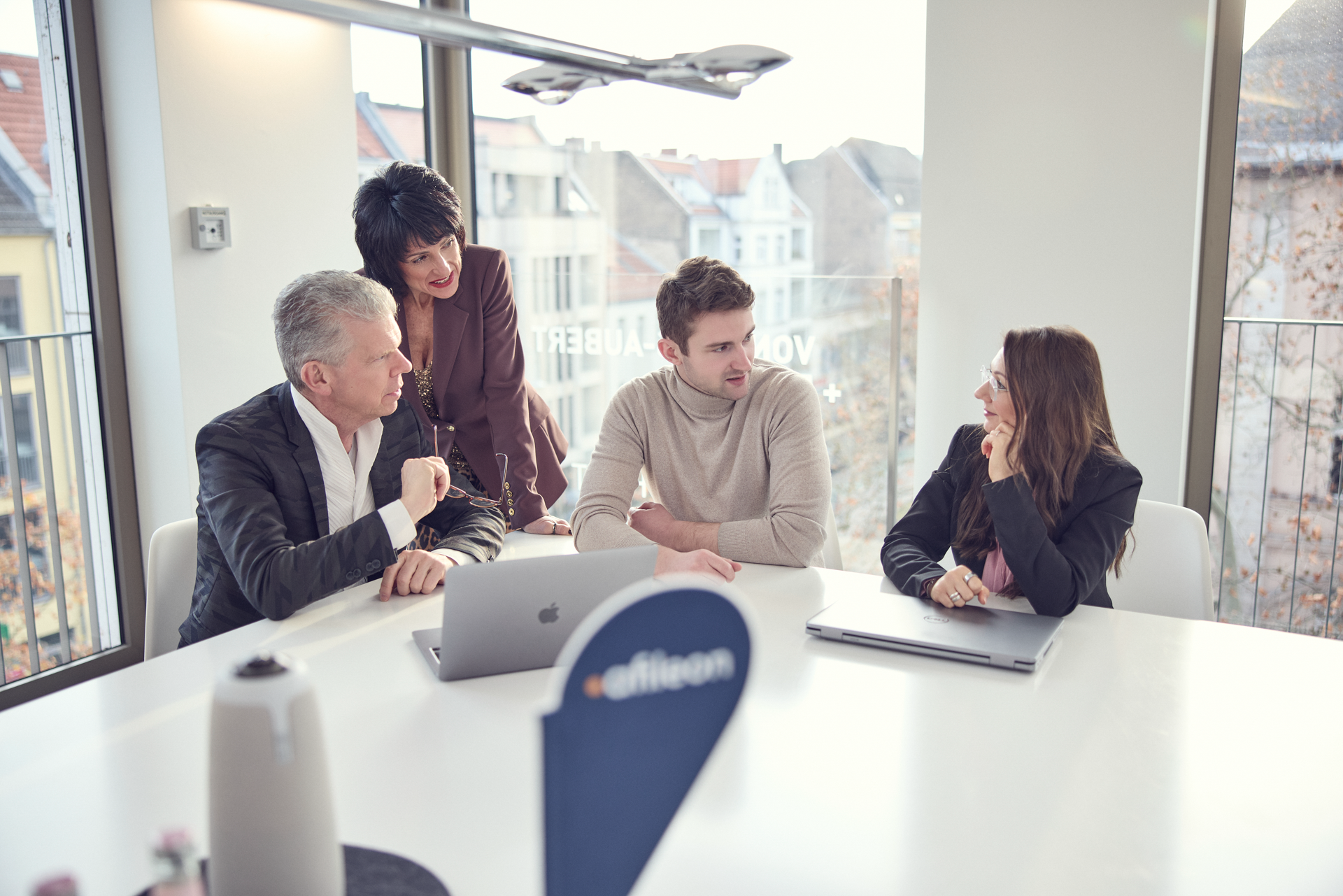 Vier Personen in einem hellen Besprechungsraum diskutieren um einen Tisch, während ein Laptop geöffnet ist. Zwei Männer und zwei Frauen sind anwesend, wobei eine Frau auf einen der Männer zeigt. Im Hintergrund sind Fenster mit Blick auf eine Stadtlandschaft zu sehen.