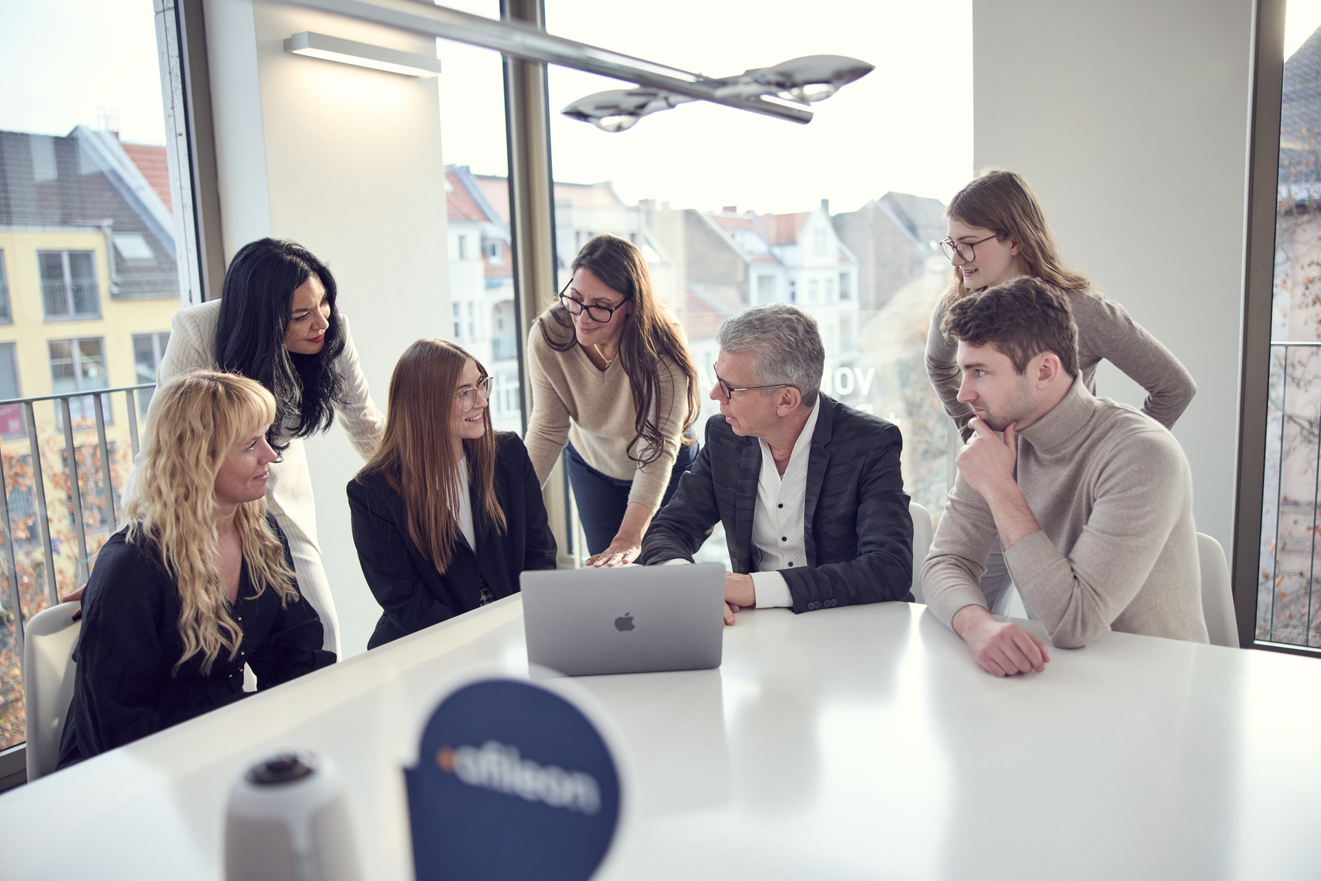 Eine Gruppe von sieben Personen versammelt sich um einen Tisch mit einem Laptop in einem modernen Büroraum, während sie zusammen an einem Projekt arbeiten und sich gegenseitig austauschen. Im Hintergrund sind Fenster mit Blick auf eine städtische Umgebung zu sehen.