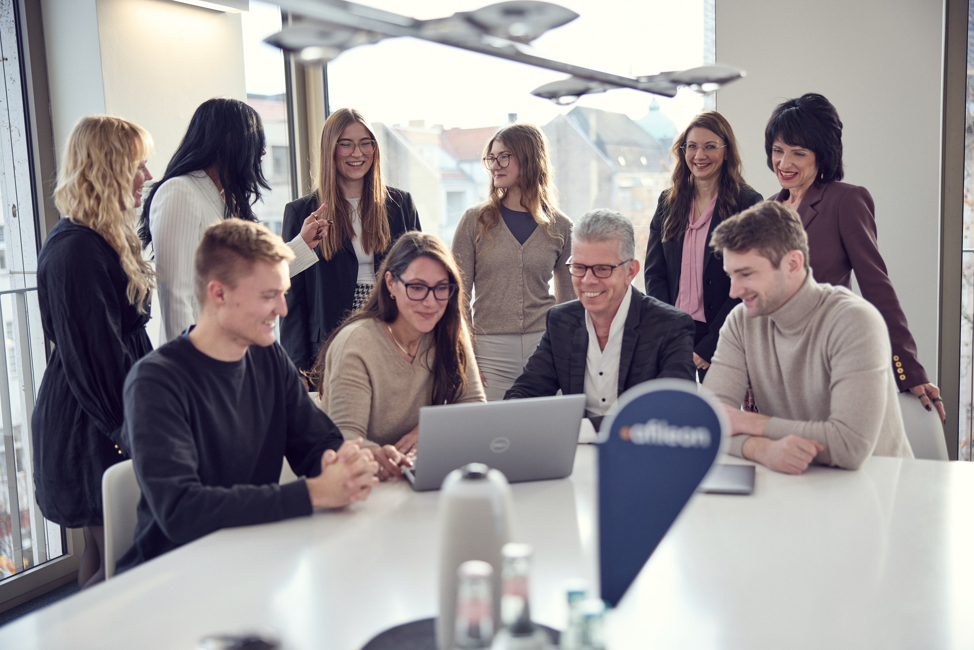Eine Gruppe von acht Personen in einem modernen Büroraum, die um einen Tisch versammelt sind und zusammen an einem Laptop arbeiten, während einige lächeln und andere aufmerksam schauen. Im Hintergrund sind Fenster mit Stadtblick zu sehen.