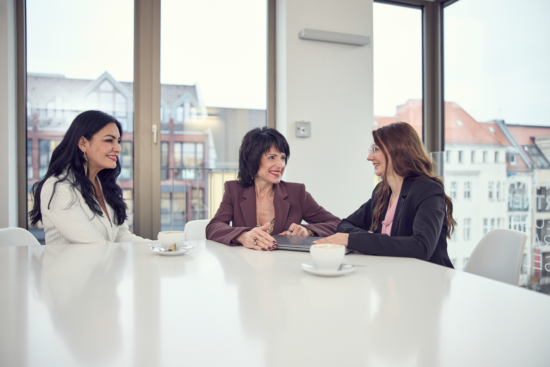 Drei Frauen sitzen an einem hellen Tisch in einem modernen Büroraum, während sie sich unterhalten. Vor ihnen stehen Kaffeetassen, und eine von ihnen hat einen Laptop geöffnet. Durch die großen Fenster ist der Blick auf städtische Architektur zu sehen.